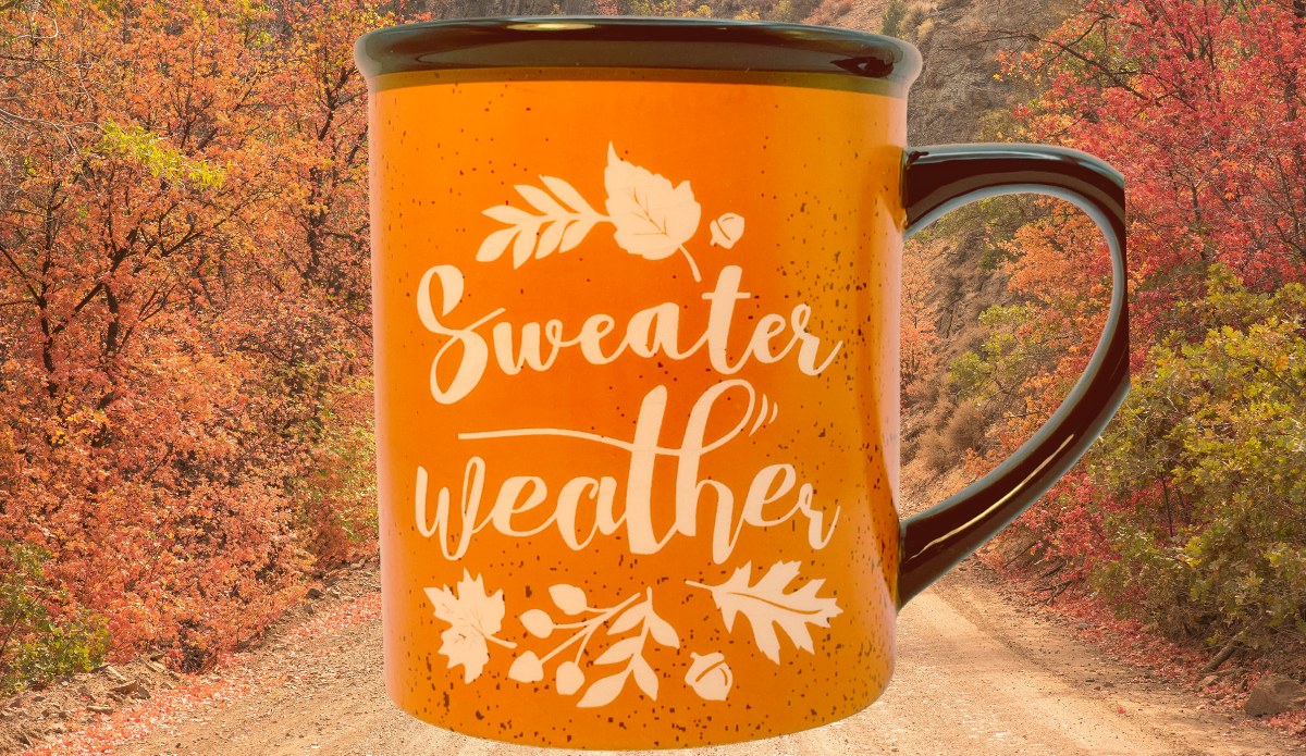 Sweater Weather Orange Mug displayed against a wooded autumn landscape with orange and red fall foliage