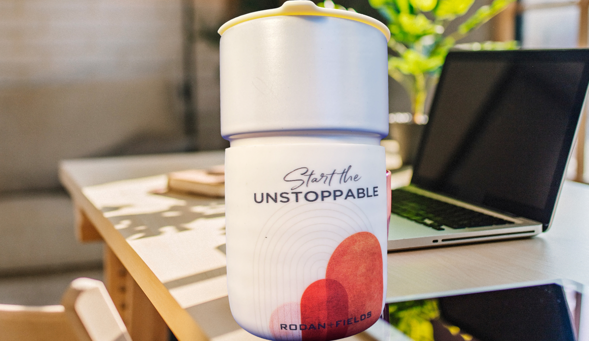 Rodan + Fields Start The Unstoppable Travel Mug on work desk beside laptop in bright motivational workspace