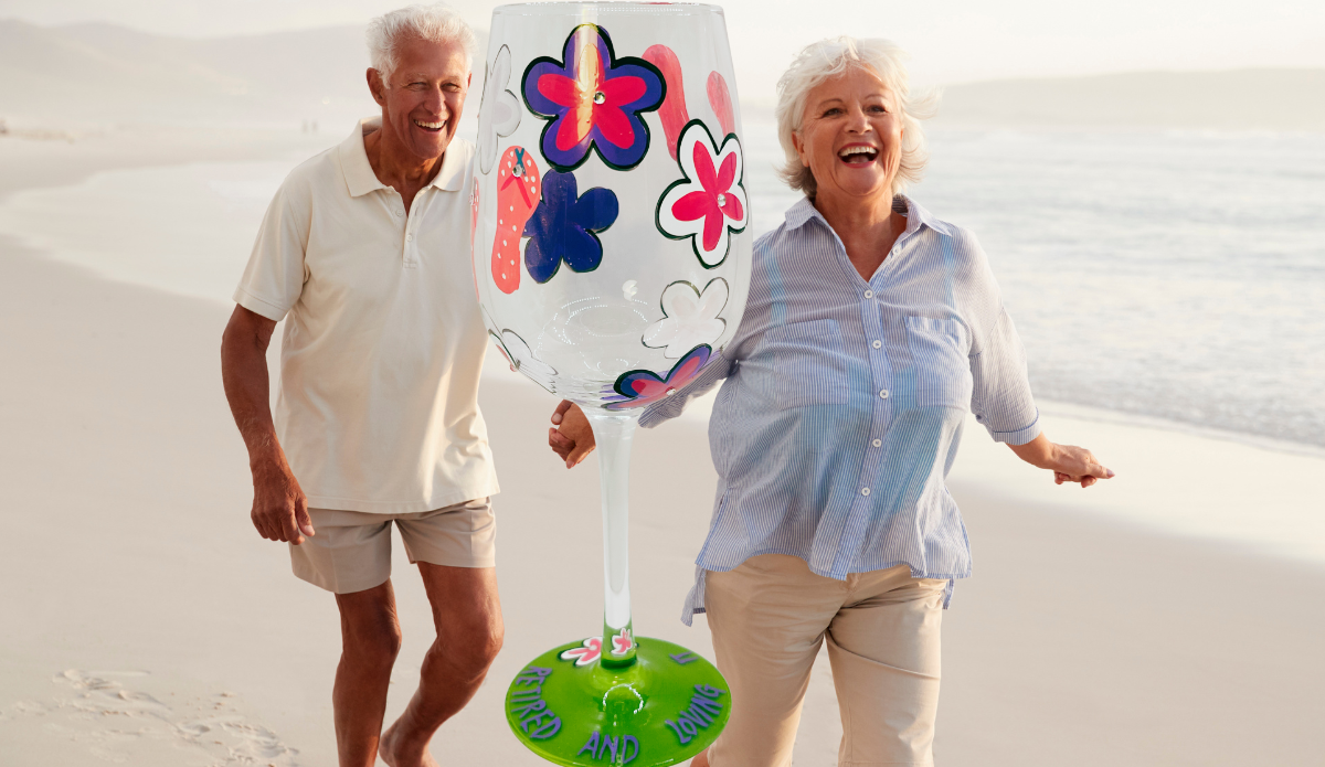 Retired And Loving It Wine Glass displayed with smiling retired couple on beach