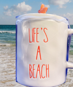 Life’s A Beach Mug displayed beside ocean waves with coral-orange starfish lid and beach backdrop
