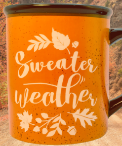 Sweater Weather Orange Mug displayed against a wooded autumn landscape with orange and red fall foliage