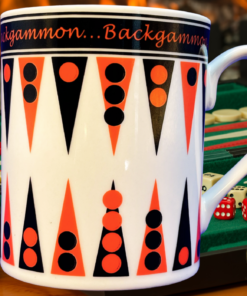 Godinger Backgammon Coffee Mug displayed beside an open green backgammon board with dice and playing pieces in a cozy fireplace setting.