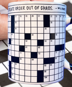 Crossword Puzzle Mug with black handle and numbered puzzle grid displayed on a large crossword page beside a brown hand.