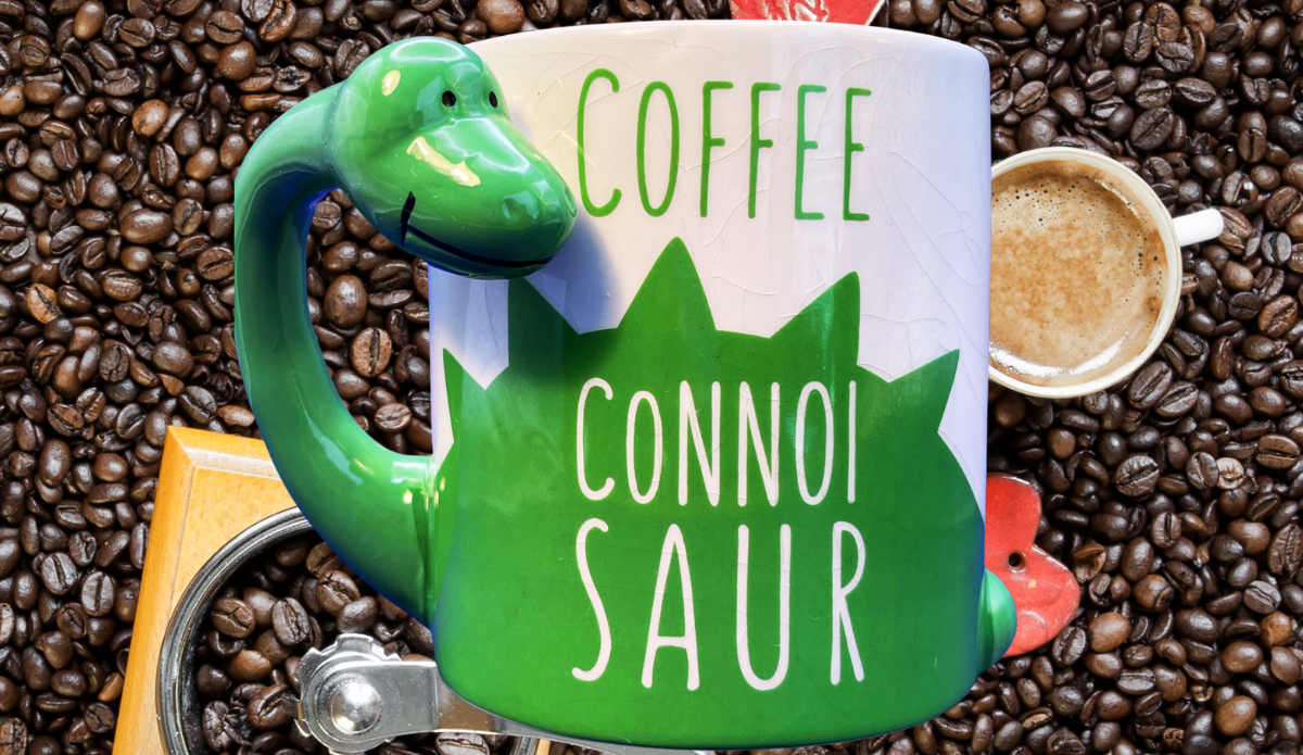 BigMouth Coffee Connoisaur Dinosaur Mug displayed beside a fresh cup of coffee and coffee beans