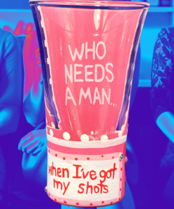 Lolita Who Needs a Man Shooter Glass displayed against blue and pink party scene with two women holding stemmed glasses