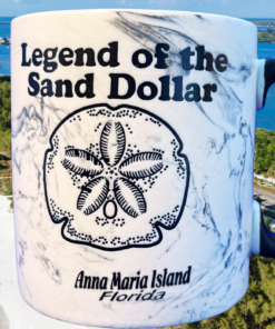 Anna Maria Island Sand Dollar Legend Mug displayed on a beach beside seashells overlooking the Gulf Coast