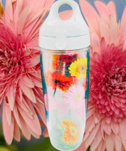 Tervis Gerbera Daisies Water Bottle With White Lid displayed against two large pink flowers with layered petals in a colorful floral lifestyle scene.
