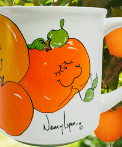 Nancy Lynn Happy Oranges Mug displayed beside fresh oranges in a citrus grove setting