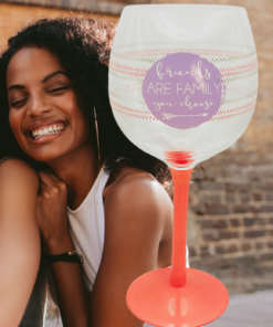 Friends Are Family Wine Glass displayed beside three smiling friends, featuring its purple friendship quote and bright coral-orange stem.