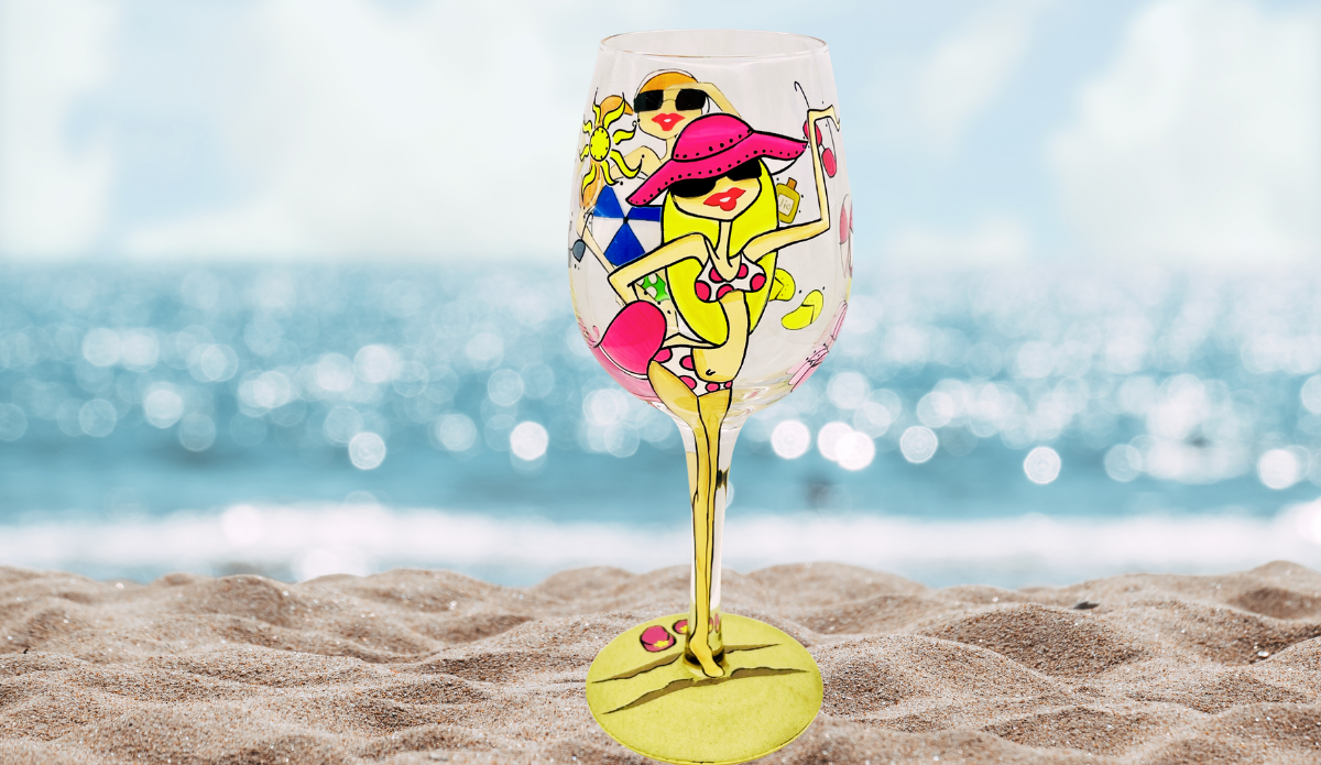 Beach Girls Hand Painted Wine Glass displayed on sand with sparkling ocean water in the background