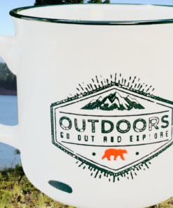 Adventure Is Out There Camping Mug displayed beside a campsite overlooking a lake and mountains