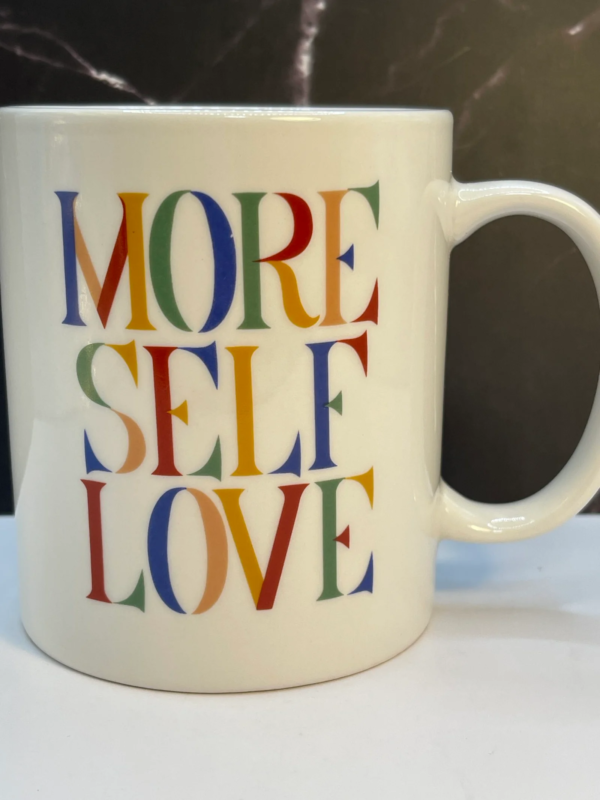 This More Self Love Mug 10-ounce white ceramic mug features bold, uplifting typography in deep blue, yellow, and red letters that proudly declare: More Self Love.