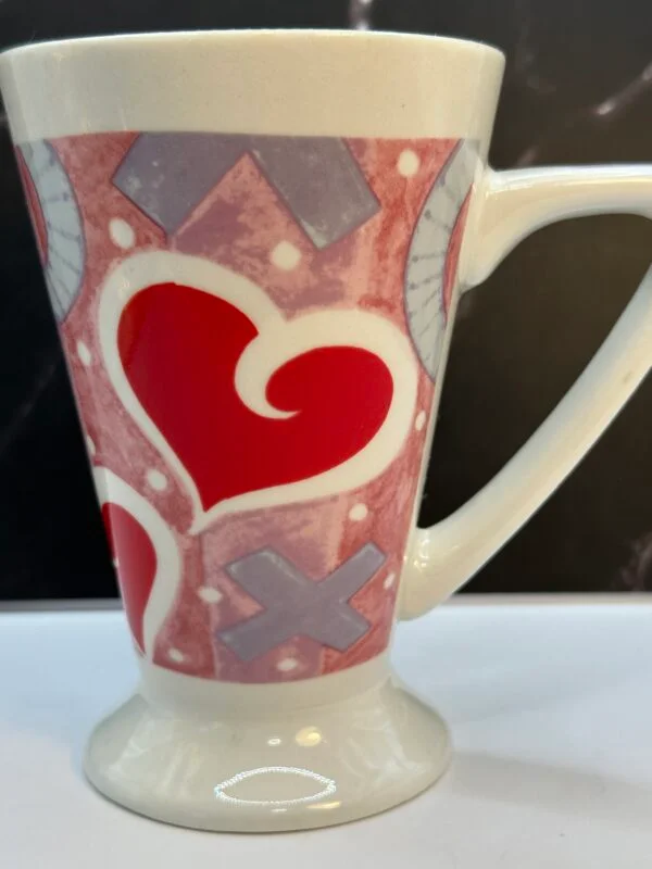 This 12-ounce white ceramic Royal Norfolk Valentine Hearts Latte Mug combines a timeless pedestal base with a vibrant and festive design. The centerpiece is a patterned pink background filled with bold red hearts. Playful Xs and Os, and dainty white polka dots that extend beautifully down the handle.