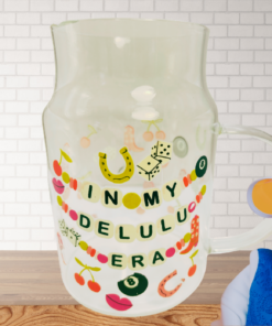 In My Delulu Era Funny Glass Mug styled on a kitchen counter with playful lucky charm graphics and a smiling older woman wearing a visor and oversized glasses beside the mug