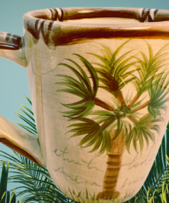Tabletops Lifestyles Bora Bora Palm Tree Hand Painted Mug displayed among lush tropical palm leaves with a peaceful island atmosphere