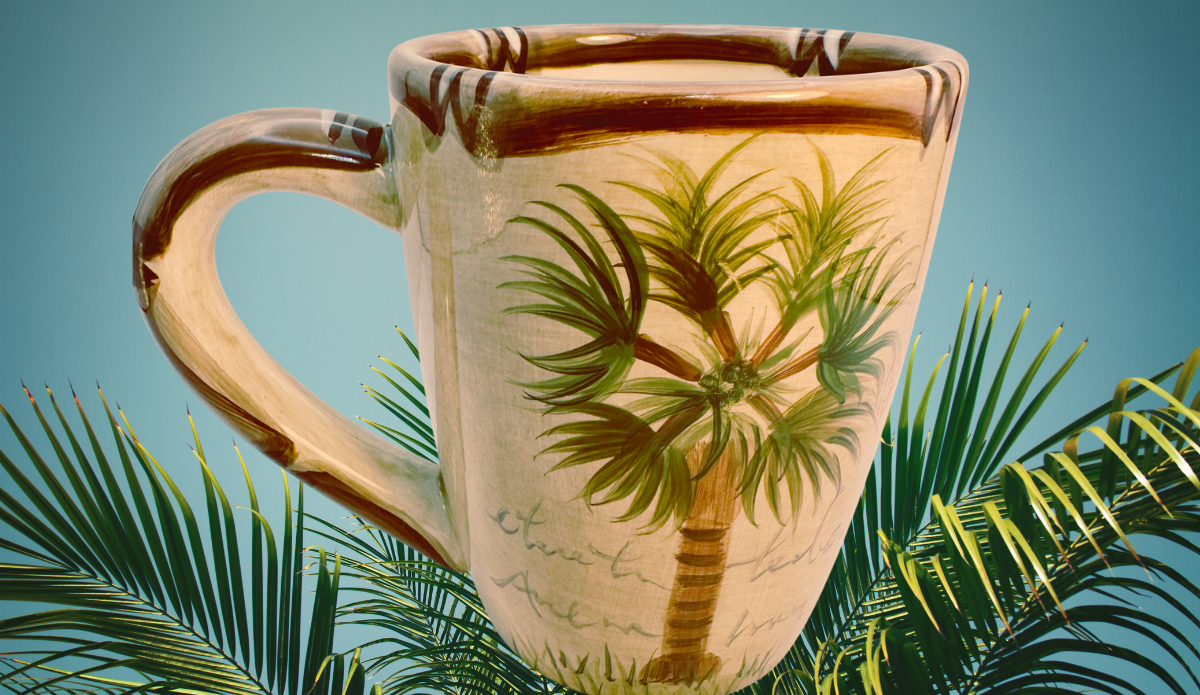 Tabletops Lifestyles Bora Bora Palm Tree Hand Painted Mug displayed among lush tropical palm leaves with a peaceful island atmosphere