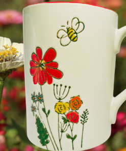 TAG Etched Flowers Bumble Bee Mug displayed in a colorful flower garden beside a bee resting on a white blossom