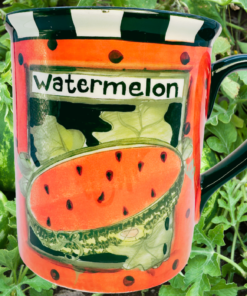 Susan Winget Watermelon Seed Packet Coffee Mug displayed among green watermelon vines with two whole watermelons in a garden setting.