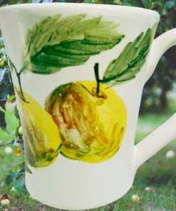 Susan Rubin Golden Apple Mug displayed in an apple orchard with hand-painted golden fruit, green leaves, flared rim, and white handle.