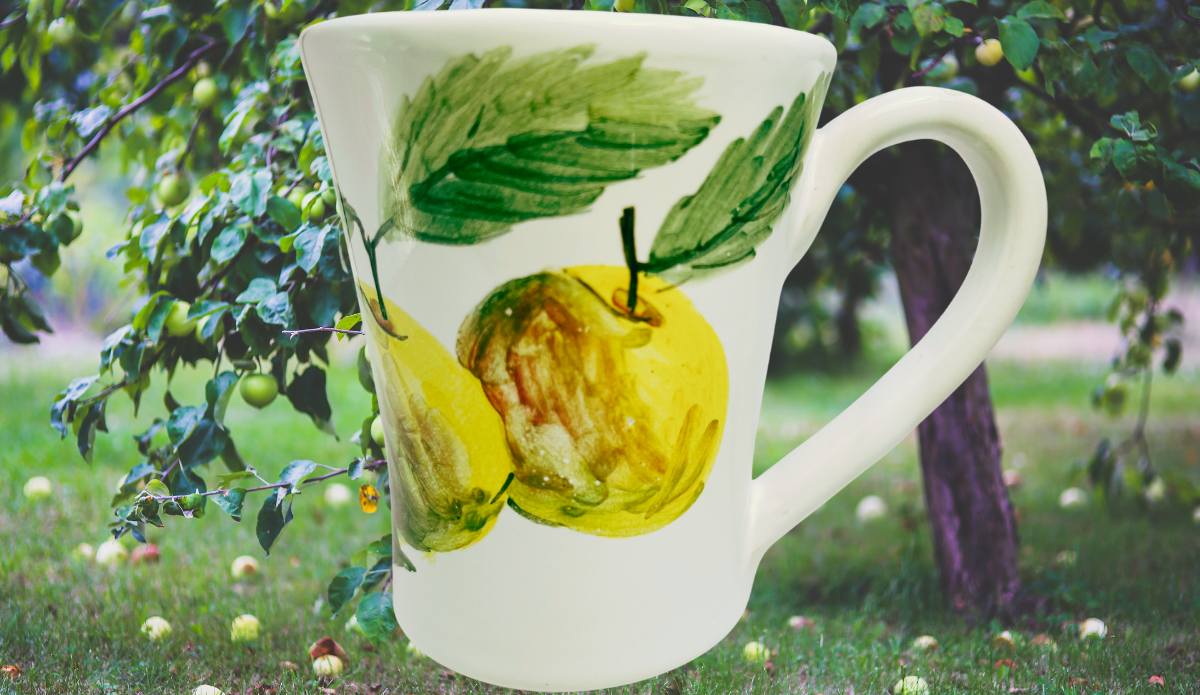 Susan Rubin Golden Apple Mug displayed in an apple orchard with hand-painted golden fruit, green leaves, flared rim, and white handle.