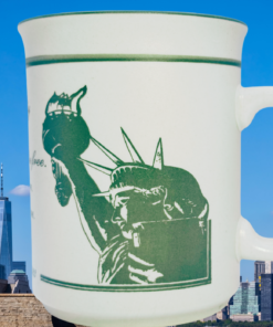 Statue of Liberty Emma Lazarus Mug displayed beside Lady Liberty and the New York City skyline under a blue sky