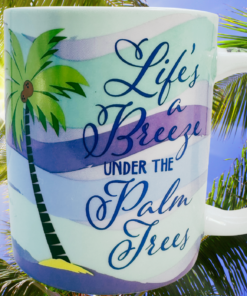 Cape Shore Life's A Breeze Under The Palm Trees Mug displayed among lush tropical palm fronds outdoors