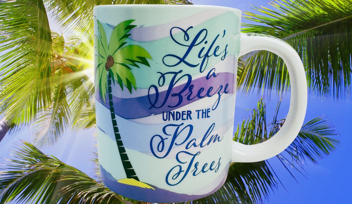 Cape Shore Life's A Breeze Under The Palm Trees Mug displayed among lush tropical palm fronds outdoors