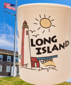 Mary Ellis Long Island Lighthouse Mug displayed beside a coastal lighthouse beneath a bright blue New York sky