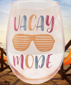 Vacay Mode Stemless Wine Glass displayed on a sandy beach beside two beach chairs overlooking the ocean