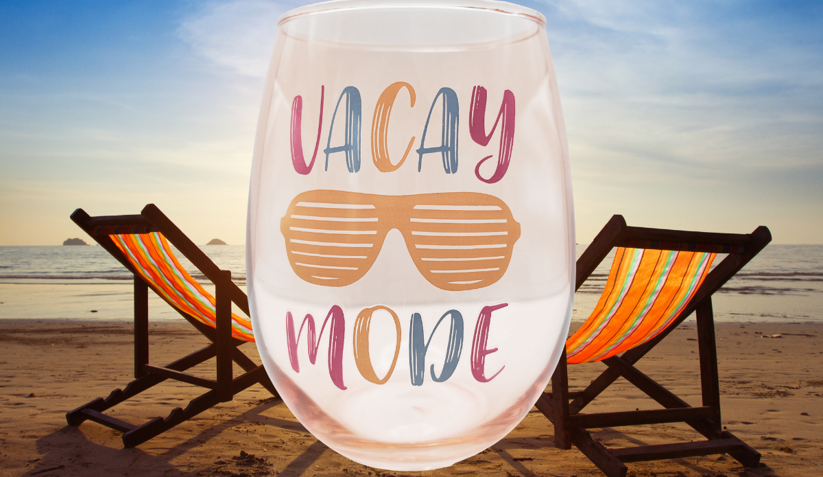 Vacay Mode Stemless Wine Glass displayed on a sandy beach beside two beach chairs overlooking the ocean