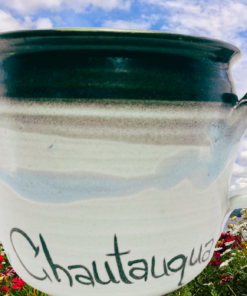 Everett Lake House Chautauqua Pottery Mug displayed in a colorful lakeside flower garden beneath a cloudy blue sky
