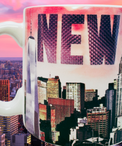 City Merchandise Pink New York Skyline Mug displayed against a colorful Manhattan sunset skyline