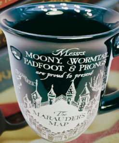 Lifestyle scene featuring the Harry Potter Marauder's Map ceramic mug displayed with Harry Potter books, eyeglasses, and a cozy striped blanket, highlighting the glossy black finish and detailed Marauder's Map artwork.