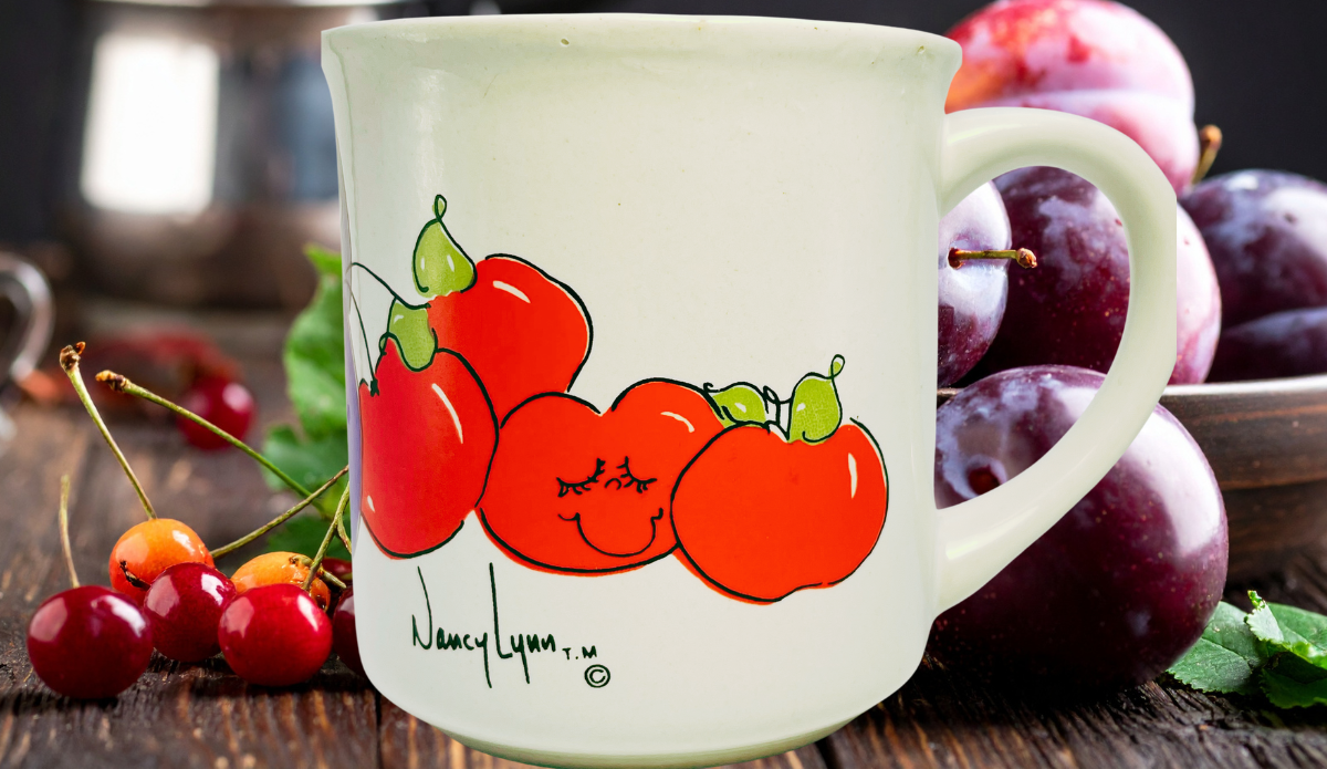 Nancy Lynn Plums and Cherries Mug displayed with ripe purple plums, red cherries, green leaves, and smiling fruit artwork.