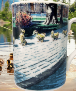 Ducklings II Mug displayed beside a peaceful pond with ducklings swimming among green reeds and natural scenery.