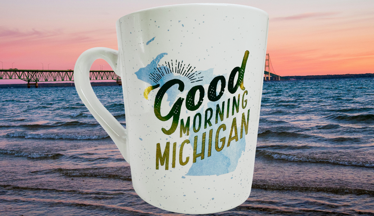 Good Morning Michigan Coffee Mug displayed beside blue water and a Michigan bridge at sunset