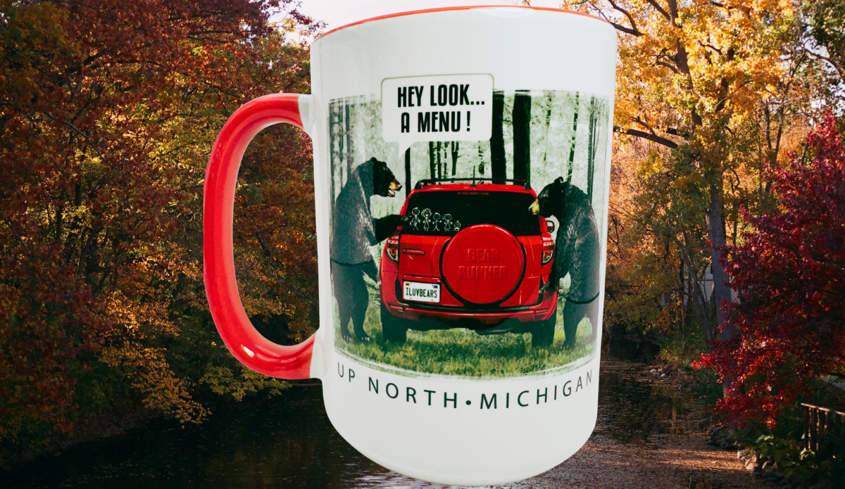 Up North Michigan Black Bear Funny Coffee Mug displayed against colorful autumn woods beside water