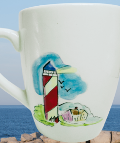 Watercolor Lighthouse Coffee Mug displayed beside ocean and red roof lighthouse