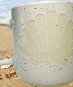 Speckled Scallop Shell Mug displayed on sandy beach with seashells and ocean water