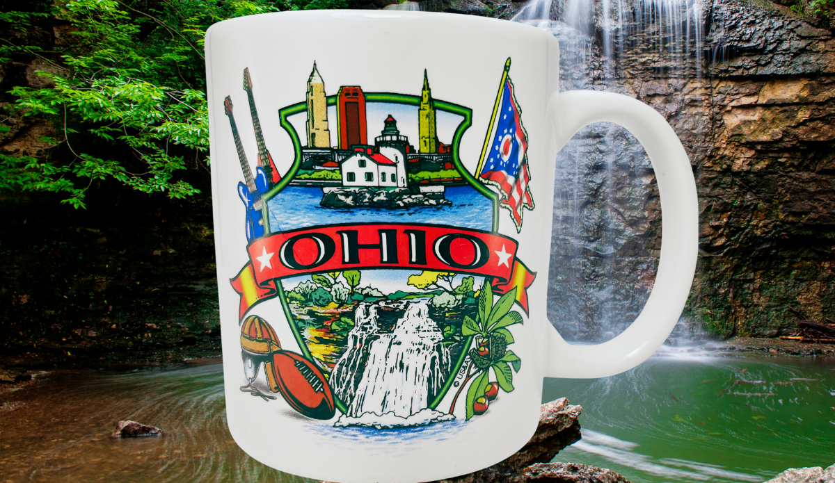 Famous Ohio Sights Souvenir Mug displayed at a wooded waterfall with colorful Ohio skyline, flag, guitars, and football design