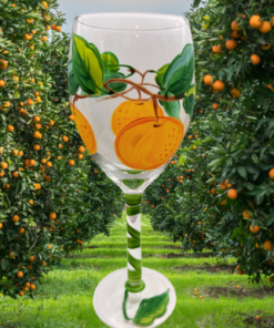 Hand Painted Oranges Wine Glass displayed in an orange grove with citrus trees, green foliage, and ripe oranges