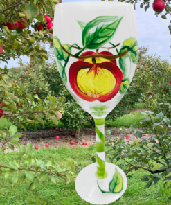 Hand Painted Apples Wine Glass displayed in an apple orchard with red apples and green foliage for a fall lifestyle scene
