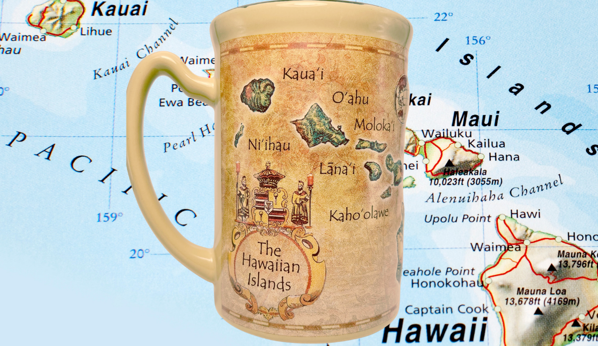 Island Heritage Hawaii Map Mug photographed against a colorful geographical map of Hawaii with blue Pacific Ocean background.