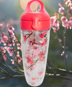 Tervis Japanese Cherry Blossom Water Bottle displayed among sunlit pink flowering branches with coral-pink lid and clear floral insulated body.