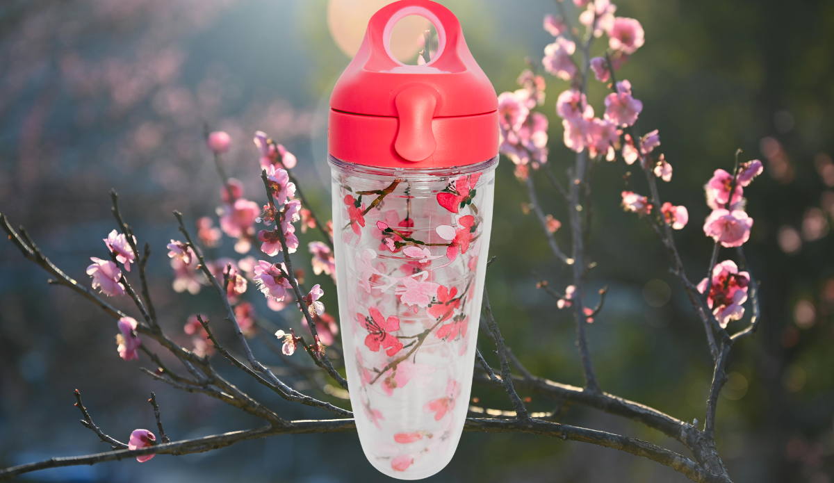 Tervis Japanese Cherry Blossom Water Bottle displayed among sunlit pink flowering branches with coral-pink lid and clear floral insulated body.