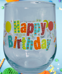 Happy Birthday Stemless Wine Glass displayed among colorful birthday candles, curling ribbons, paper decorations, and confetti.