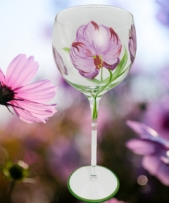 Hand Painted Purple Pink Flower Balloon Wine Glass displayed in a soft-focus garden setting with pink cosmos flowers and lavender-purple blooms.
