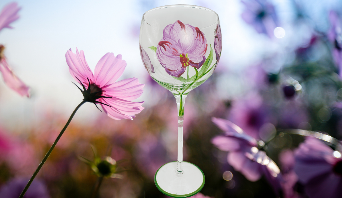 Hand Painted Purple Pink Flower Balloon Wine Glass displayed in a soft-focus garden setting with pink cosmos flowers and lavender-purple blooms.