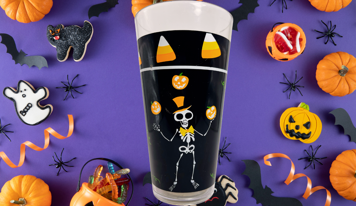 Juggling Skeleton Halloween Acrylic Tumbler surrounded by pumpkins, bats, spiders, candy, and spooky treats on purple background