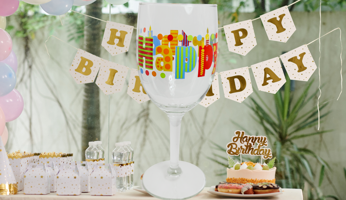 Happy Birthday Colorful Lettering Wine Glass displayed at a decorated party table with birthday banner, pastel balloons, cake, and treats.